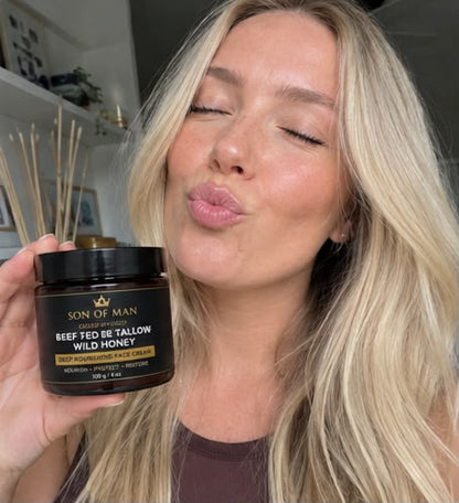 A woman with blonde hair holds up a jar of Son of Man Beef Fed Be Tallow Wild Honey deep moisturizing face cream while puckering her lips. The background is a softly blurred indoor setting.