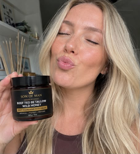 A woman with blonde hair holds up a jar of Son of Man Beef Fed Be Tallow Wild Honey deep moisturizing face cream while puckering her lips. The background is a softly blurred indoor setting.
